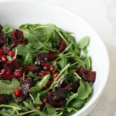 Arugula salad in a white bowl with pomegranate seeds and roasted beets.