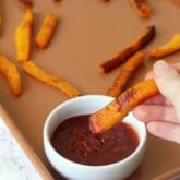 A sheet pan of butternut squash fries with one being dipped into ketchup.