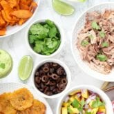 White bowls of ingredients including plantains, carnitas, olives, salsa, cilantro, and lime to make nachos