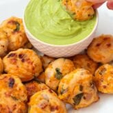 Many Mexican chicken poppers around a white cup filled with guacamole and one chicken popper dipping into it.