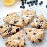 Six lemon blueberry scones on a metal cooling rack with fresh blueberries and lemon slices nearby.