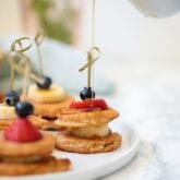 A white plate with mini pancakes on skewers with fresh fruit.