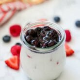 A glass jar filled with red white and blue panna cotta with fresh berries on the counter beside it.