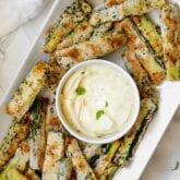 A white serving dish of zucchini fries around a white bowl of dipping sauce.