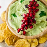 A wooden bowl of pomegranate guacamole surrounded by plantain chips.