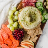 A wooden board topped with apples, carrots, pomegranates, grapes, crackers, hummus, cucumbers, and cured meats to make a charcuterie board.