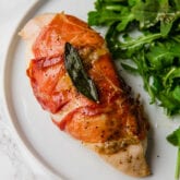 A white plate with argula salad next to a piece of proscuitto chicken.