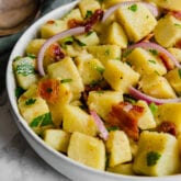 A white serving bowl filled with potato salad topped with bacon and onions.