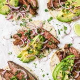 Several carne asada tacos with avocado and red onion.