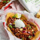 A red basket with parchment paper fille with frito pie over plantain chips with fresh lime wedges on it.