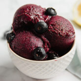 A small white bowl with blueberry lemon sorbet scoops in it and fresh blueberries.