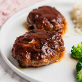 A white plate topped with japanese hamburger steaks on it with broccoli on the plate as well.