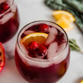 Pomegranate mocktail in a glass with ice, cranberries, and orange slices with rosemary and orange slices in the background.