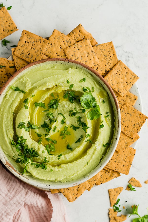 Avocado hummus in a bowl with crackers around it