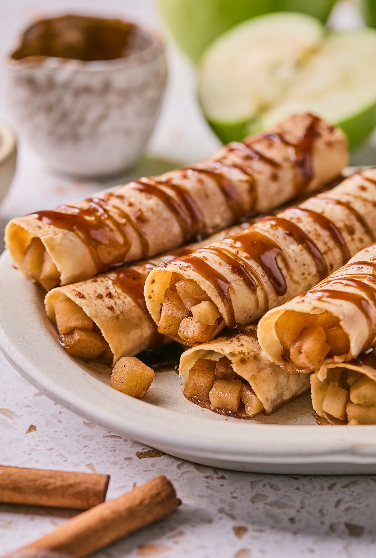 Apple pie roll ups on a plate drizzled with caramel, and with extra caramel on the side. 