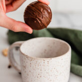 Person holding a hot cocoa bomb over a speckled mug.