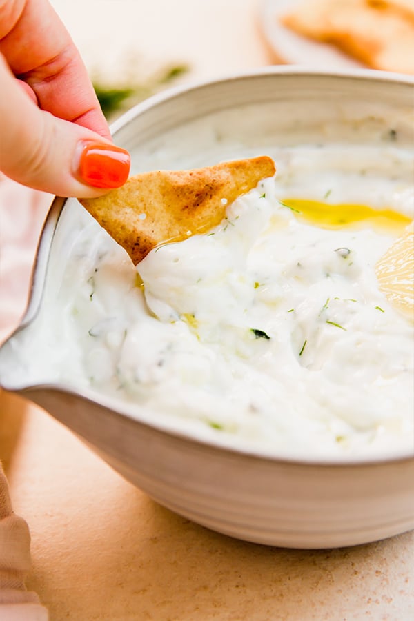 A gluten-free pita chip dipping into a bowl of dairy-free tzatziki sauce.