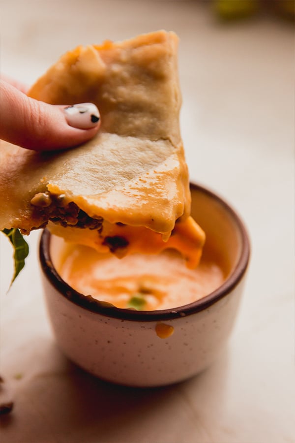 A hand dipping a baked ground beef taco into dairy-free cheese sauce.