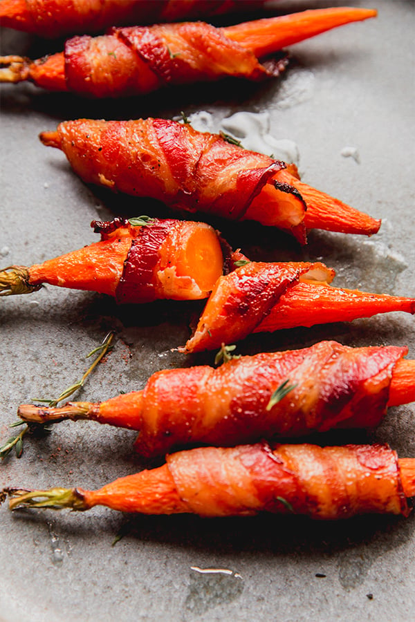 a few bacon-wrapped carrots on a plate with one cut open.