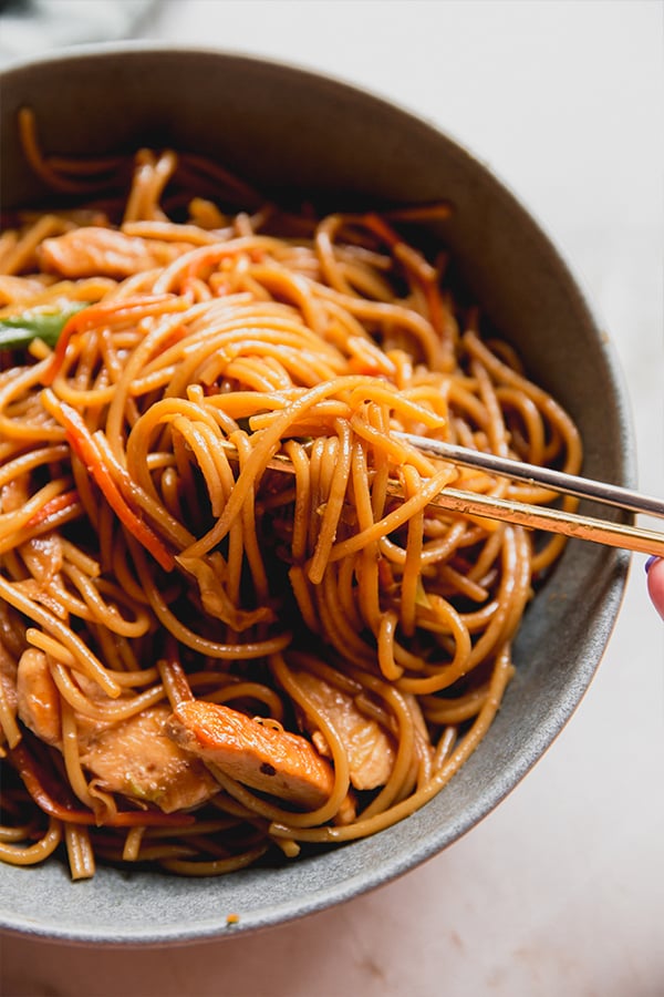 A bowl of chicken chow mein in a bowl with chop sticks taking a bite out.