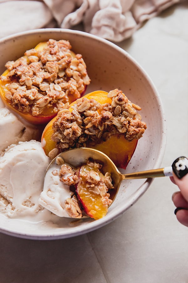 A bowl of air fryer peach crisp with dairy-free ice cream and a spoon taking a bite.