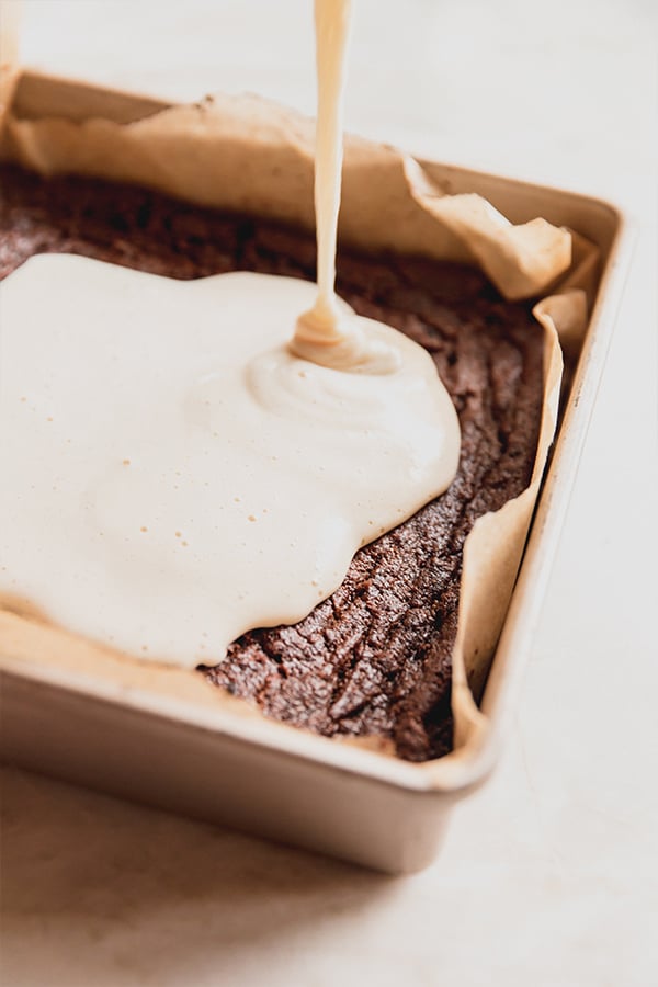 The marshmallow layer being poured over the brownie layer in a baking dish.