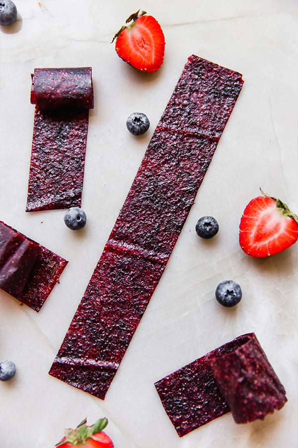 Homemade fruit roll-ups on a counter with fresh strawberries and blueberries.