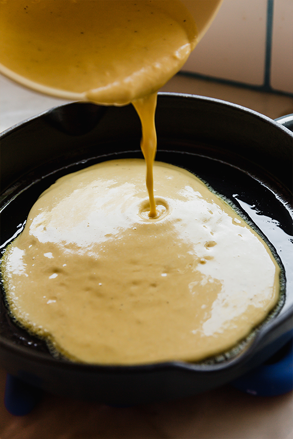 Chickpea flatbread batter being poured from a mixing bowl into a cast iron skillet to cook.