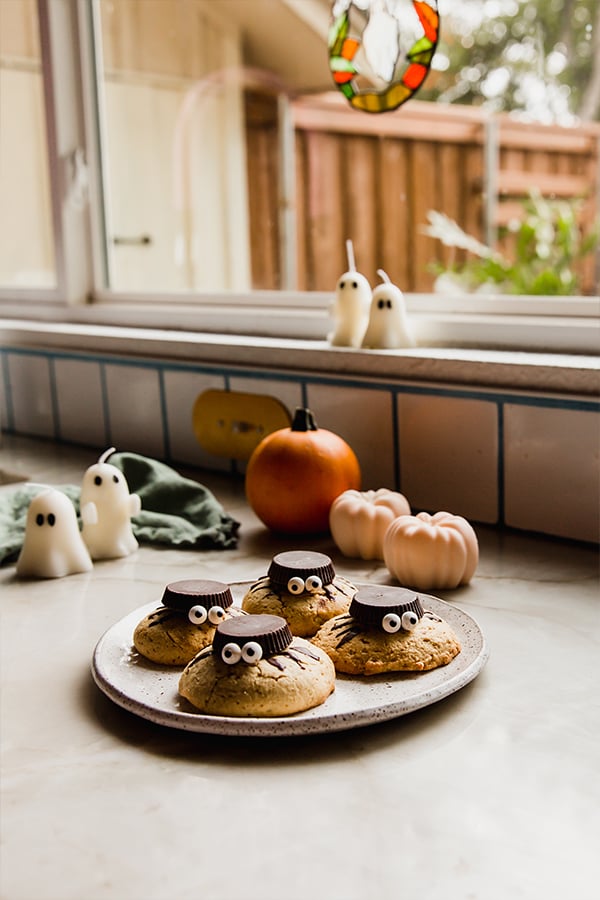 A plate of spider cookies on a plate on a counter with ghost candles and mini squashes in the background.