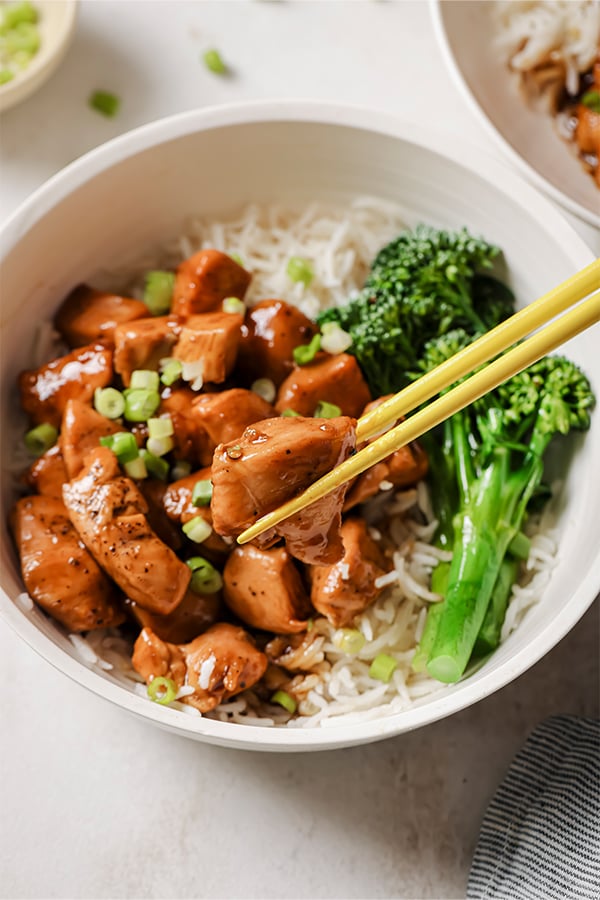 A bowl of bourbon chicken served with broccolini and rice noodles with chopsticks taking a bite.
