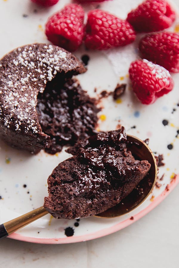 A chocolate lava cake on a plate with a spoon taking a bite out of the cake accompanied by raspberries. 