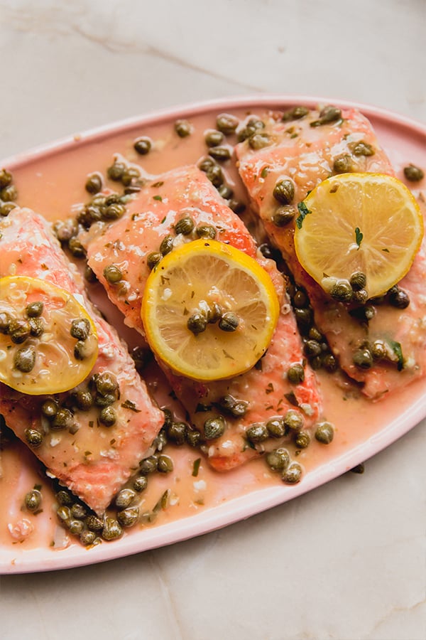 A serving platter of salmon piccata topped with capers and lemon slices.