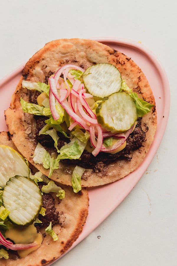 A serving plate with two smash burger tacos topped with pickled onions and pickles.