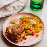 Greek lemon chicken on a pink plate with a rice and diced carrots on the side, topped with lemon wedges and a green glass with water on the side.