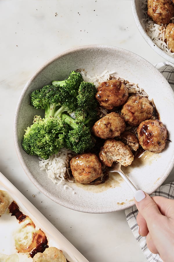 A bowl of honey garlic meatballs, broccoli, and rice with a fork take a bite out.
