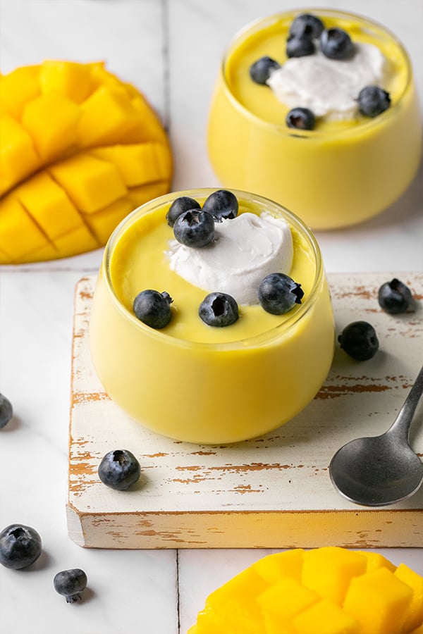 Two cups of dairy-free mango pudding topped with whipped cream and blueberries ready to be eaten.