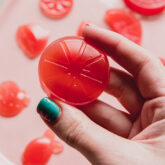 Lemon shaped pink lemonade gummy being held by a hand with blue nail polish.
