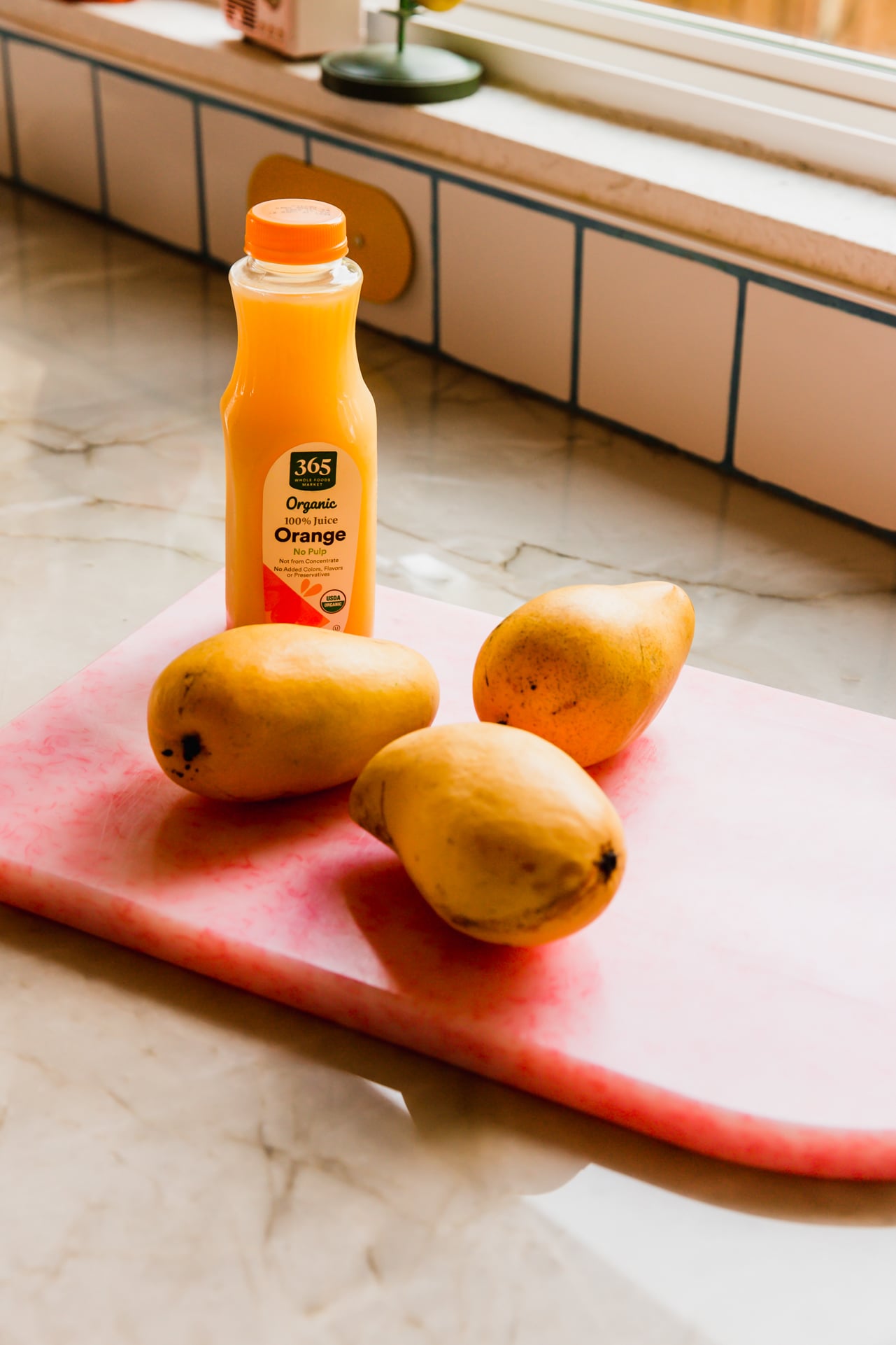 Three mangos and orange juice on a pink cutting board.