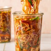 Homemade cup of noodles with ramen, veggies and broth in a glass jar with chopsticks pulling the noodles.