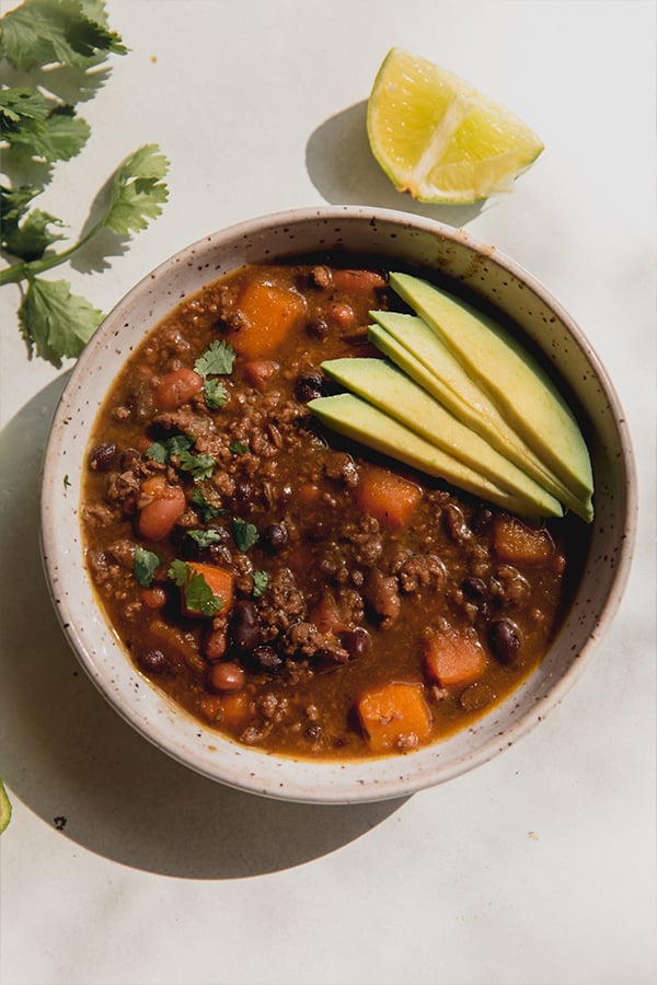 A bowl of chili topped with avocado, cilantro, and with lime wedges on the side. 