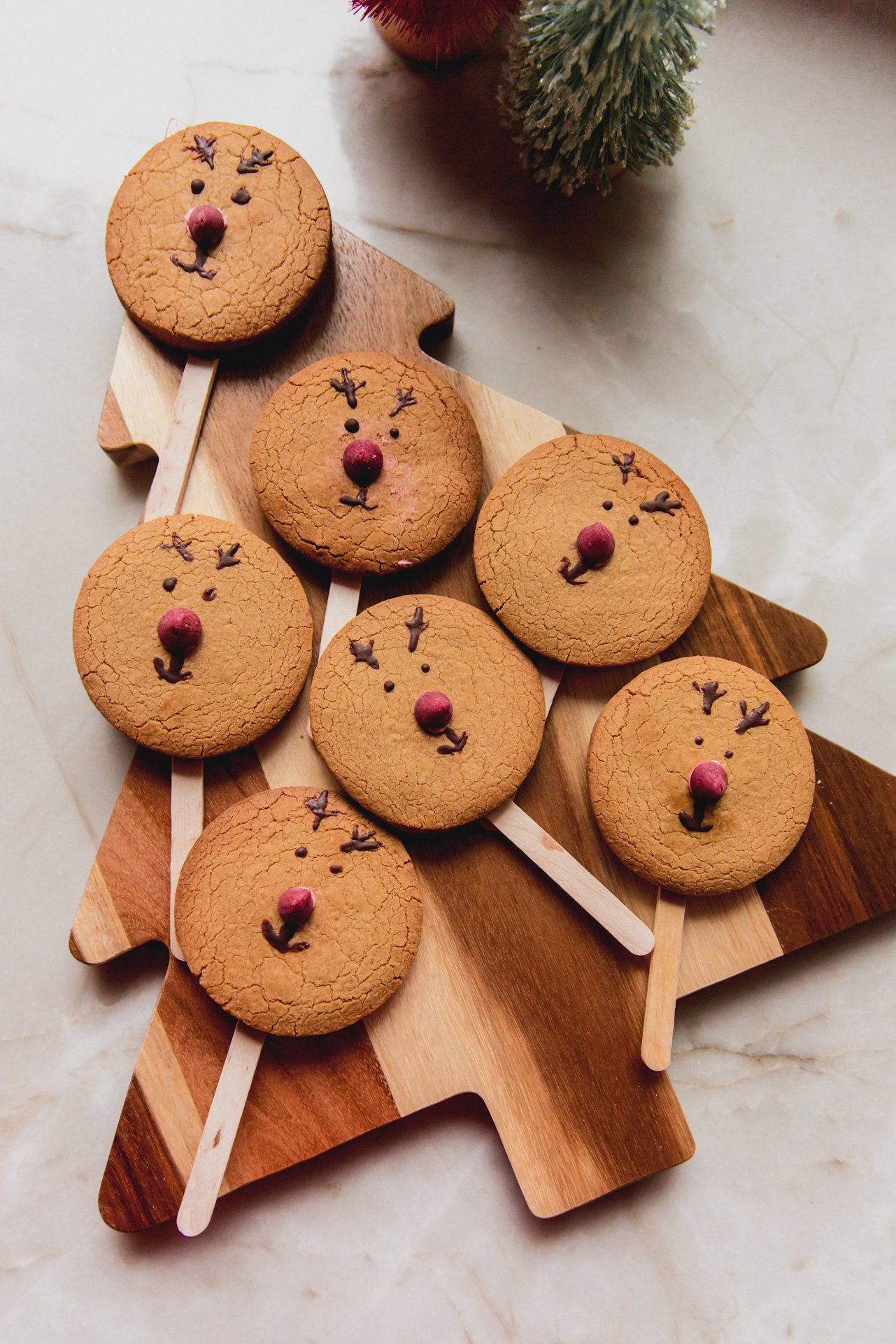 Reindeer cookies with popsicle sticks laid out on a Chirstmas tree shaped cutting board. 