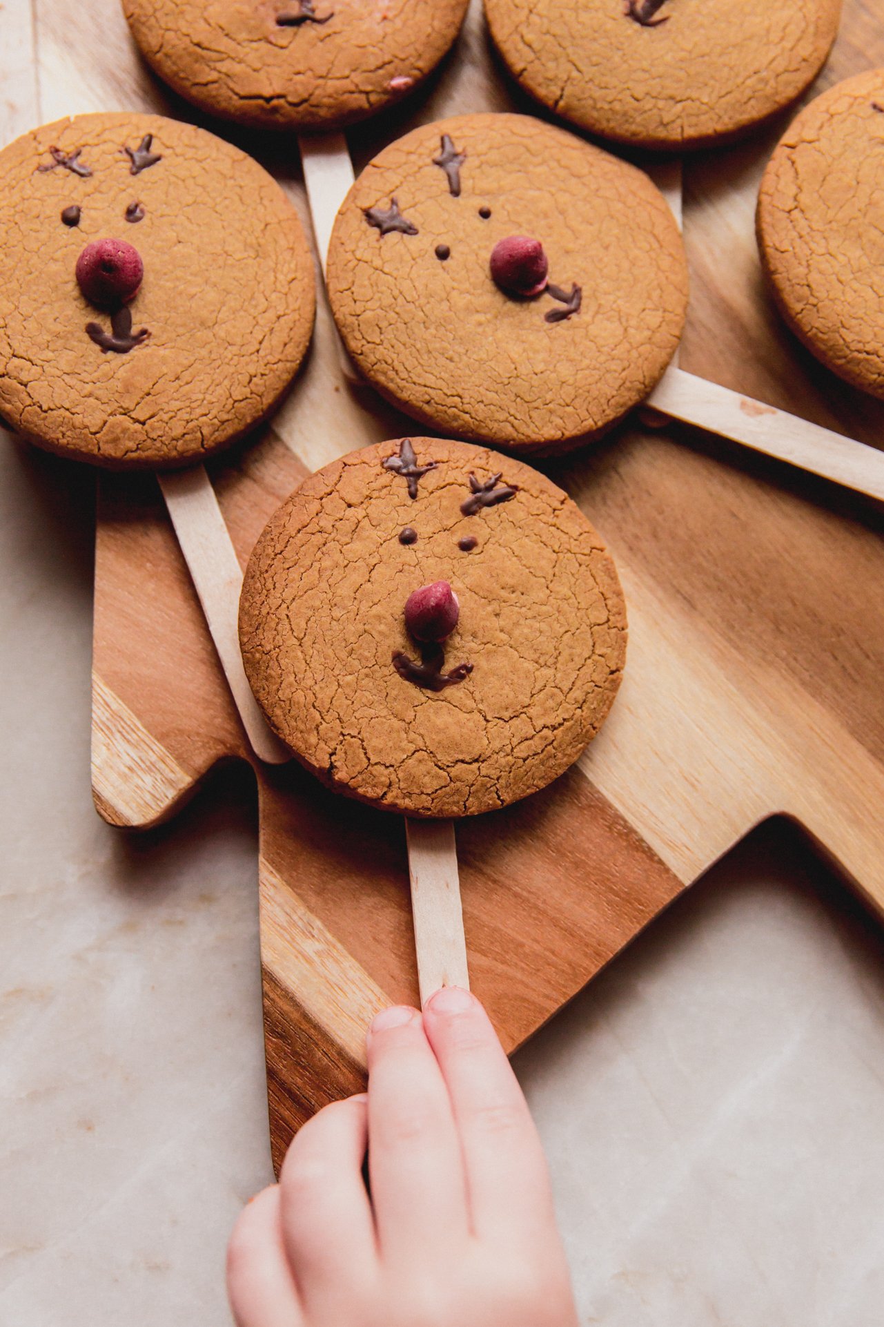 Reindeer cookies on a cutting board with a child's hand reaching from them. 