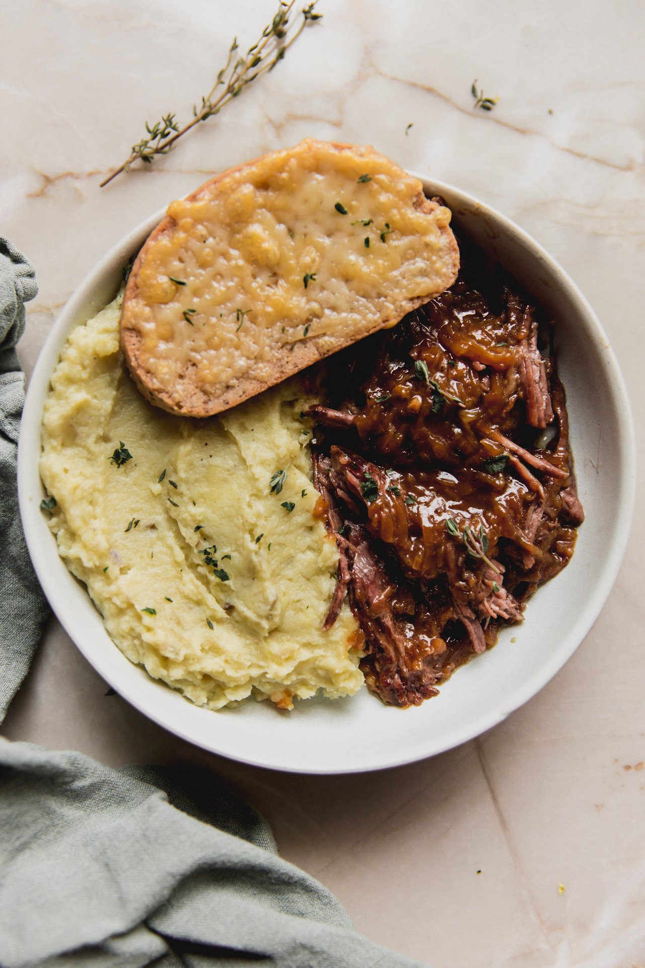 French onion pot roast in a shallow bowl with mashed sweet potato, cheese toast, and topped with herbs.