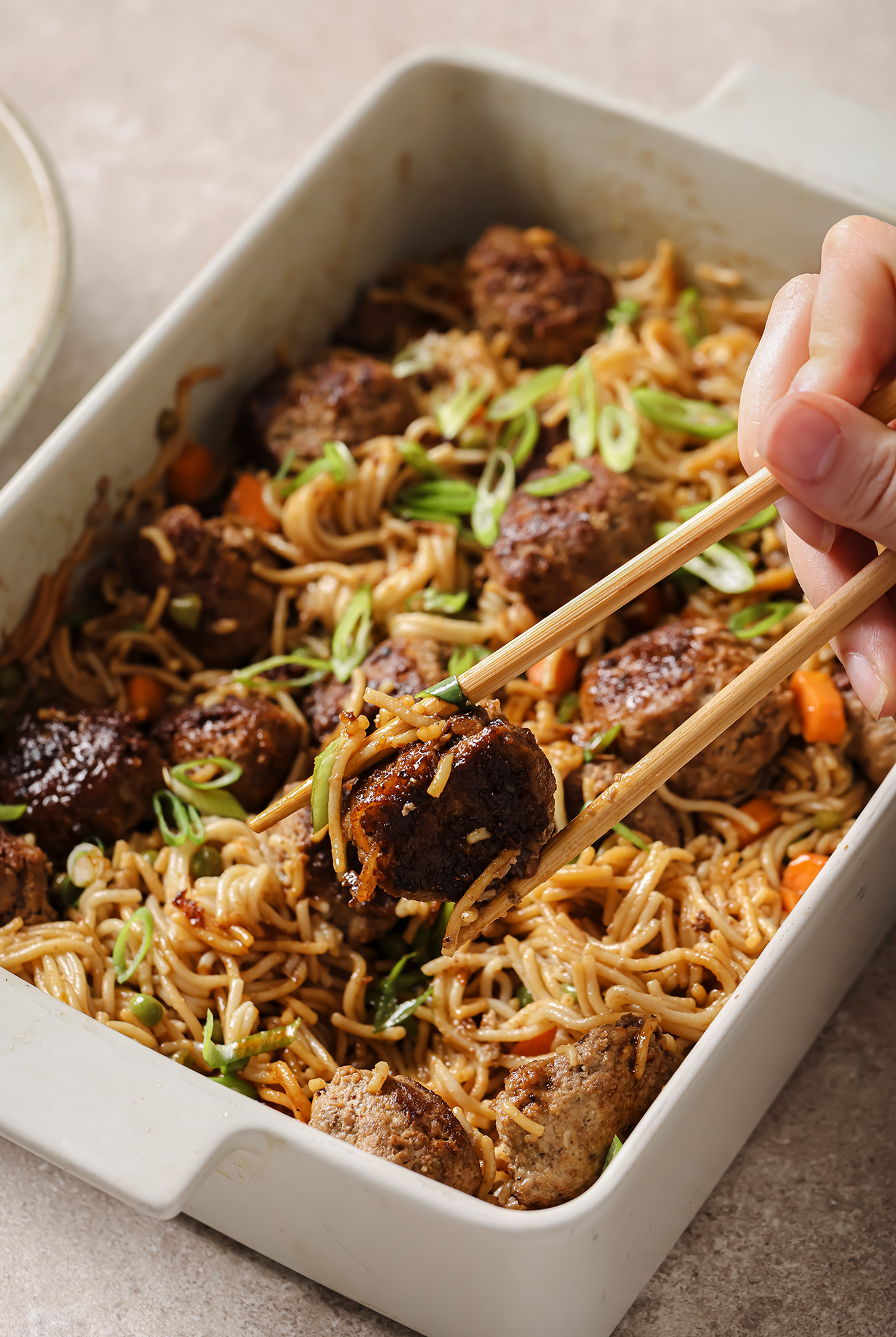 chopsticks grabbing a meatball from a casserole dish with teriyaki meatballs and ramen