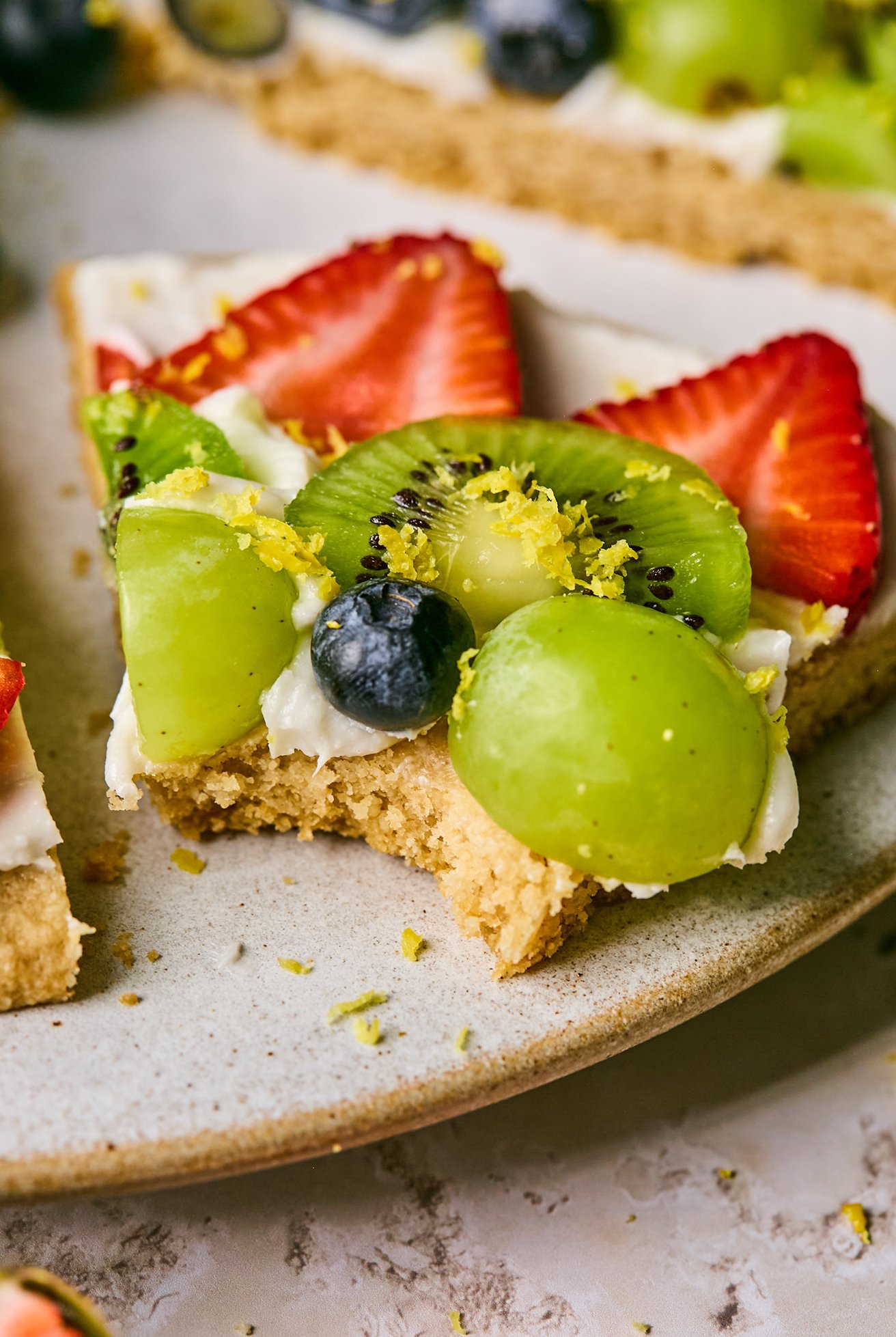 A slice of sugar cookie fruit pizza with a bite taken out of it. 