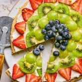 A sugar cookie fruit pizza on a plater with one slice being cut out.