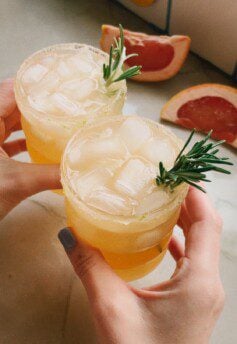 Two grapefruit palomas in hands cheersing.