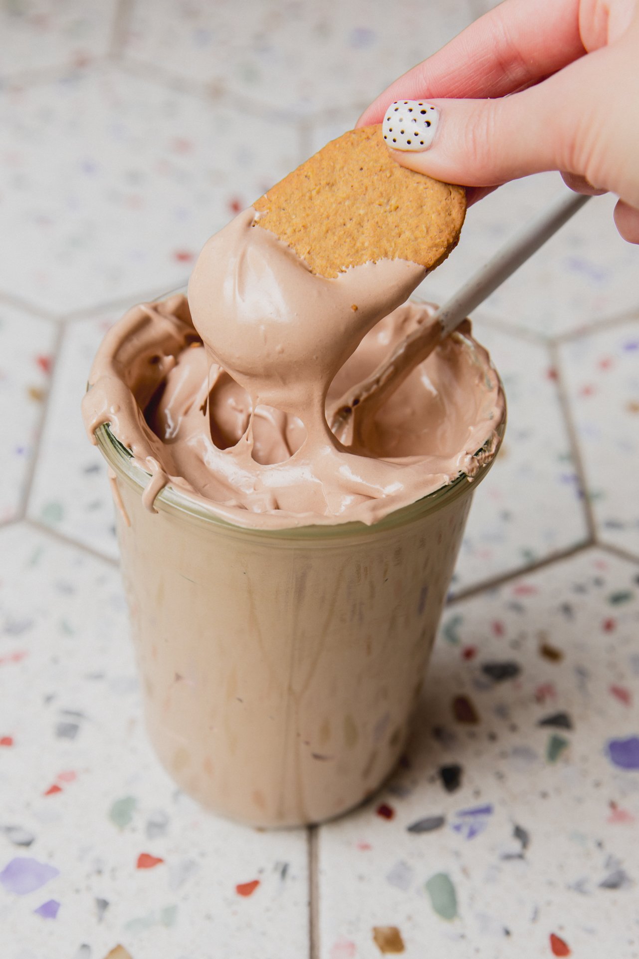 A glass container of chocolate espresso marshmallow fluff sitting on a counter with a cookie dipping into it.