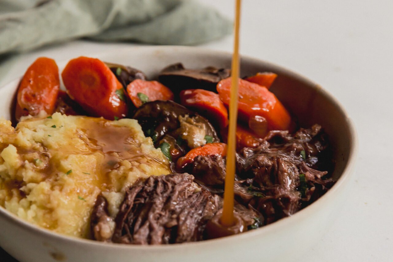 A bowl with pot roast, sweet potatoes, and vegetables with gravy being poured into it.
