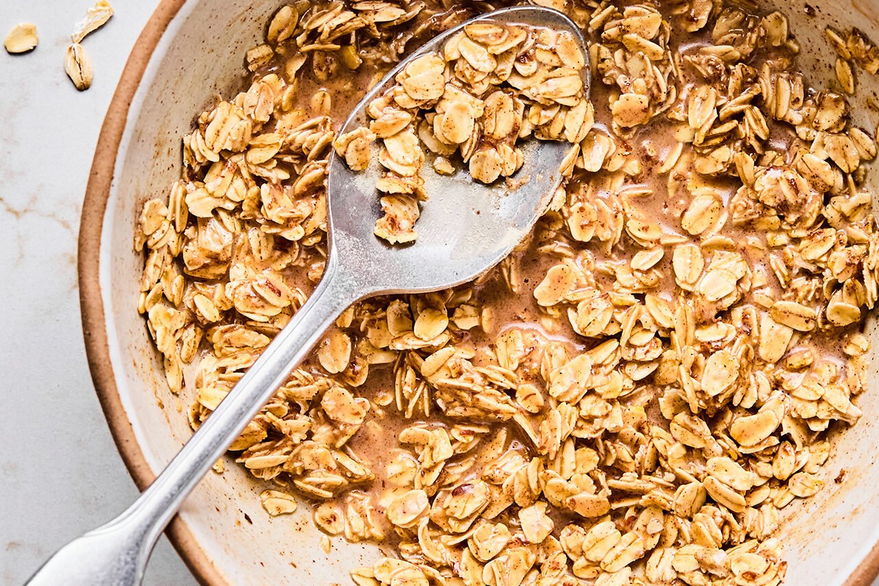 A bowl filled with the mixture of oats, protein powder, cinnamon, maple syrup, almond milk, and coconut oil after being stirred with a metal spoon.